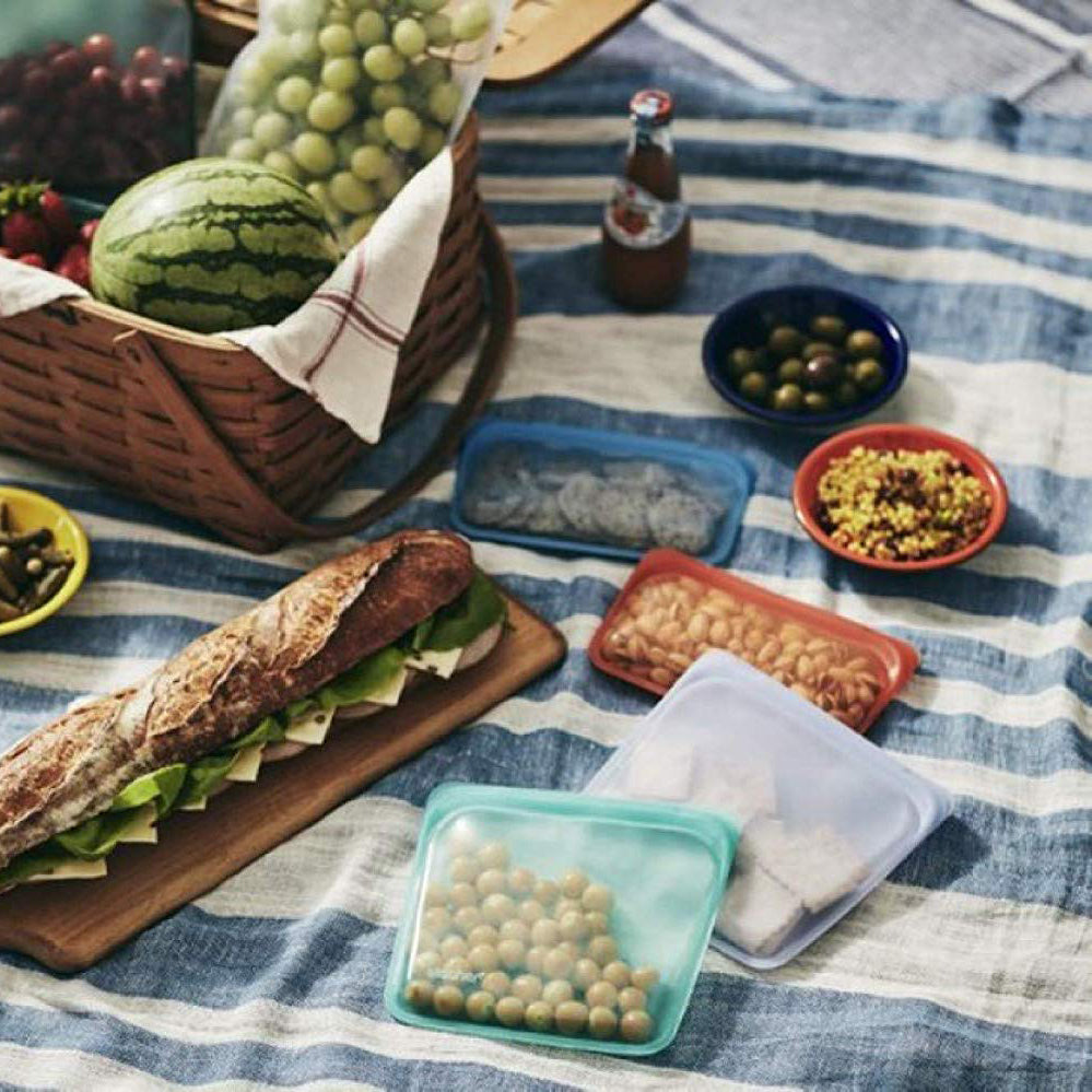 Bodegón de picnic con diferentes bolsas de silicona platino Stasher, pequeñas y medianas, de diferentes colores con diferentes aperitivos en su interior - LLevar & LLevar